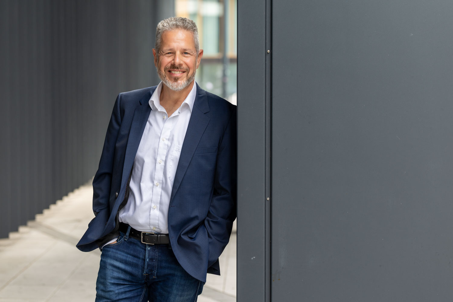 Portrait von Berater und Executive Coach für Führung und Kommunikation Jürgen Pfeiler, der mit Händen in den Hosentaschen an einen Pfeiler angelehnt freundlich lächelt.
