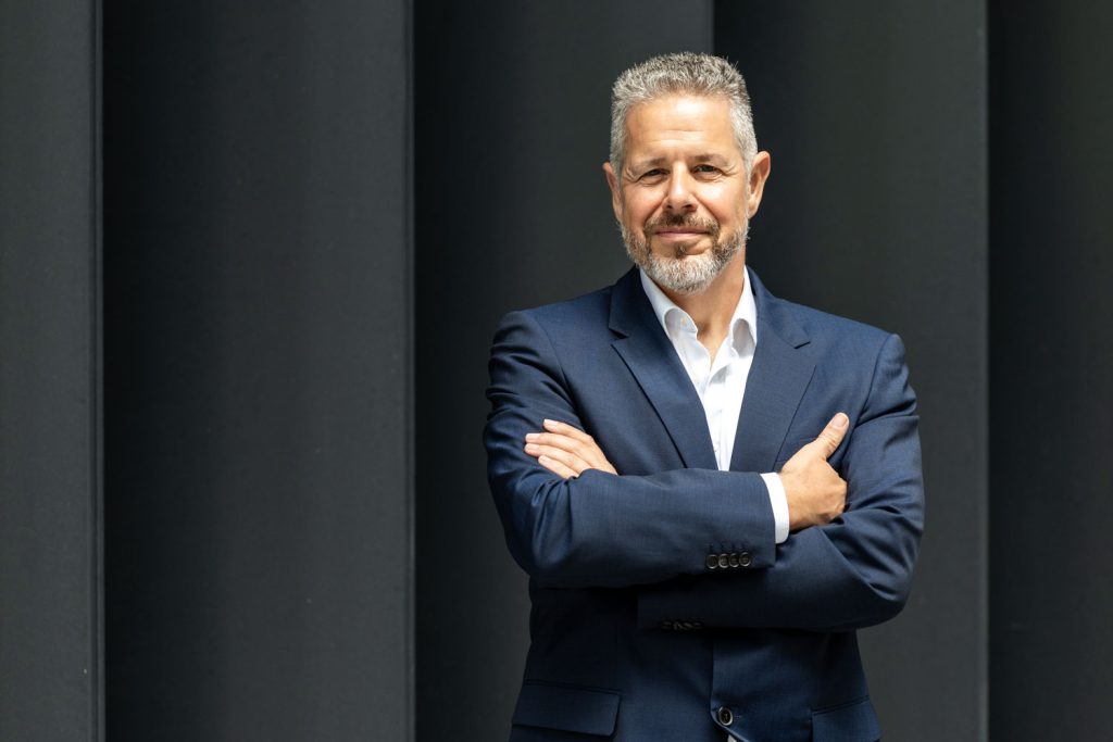 Portrait von Berater und Executive Coach für Führung und Kommunikation Jürgen Pfeiler, der mit verschränkten Armen vor einer dunkelgrauen Wand steht.