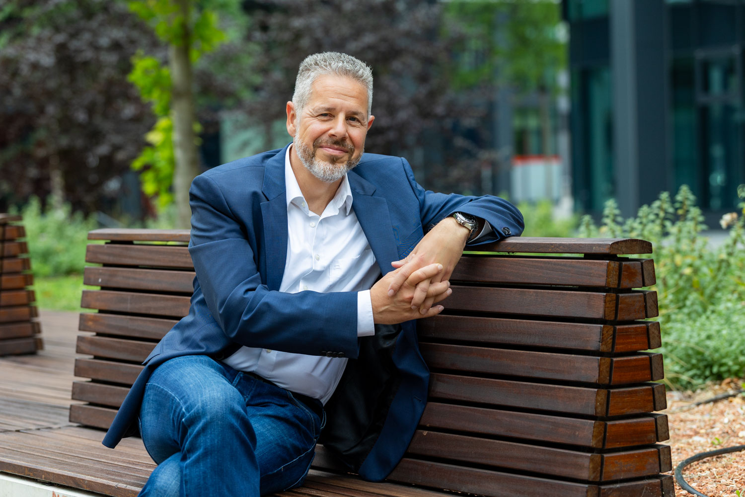 Portrait von Berater und Executive Coach für Führung und Kommunikation Jürgen Pfeiler, der auf einer Bank sitzt und freundlich lächelt.