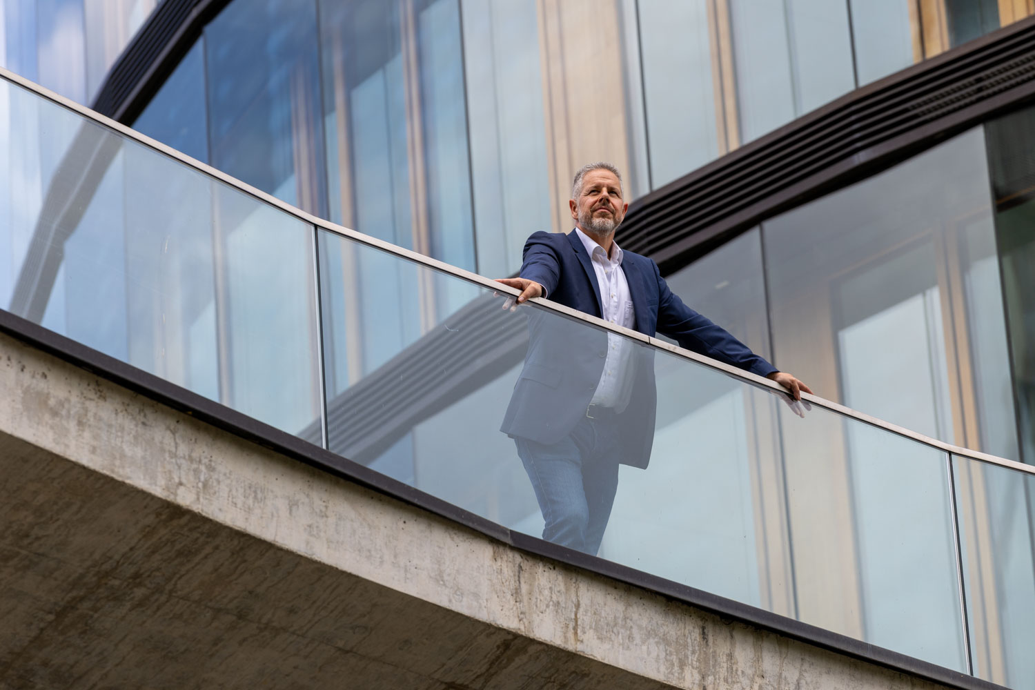 Portrait von Berater und Executive Coach für Führung und Kommunikation Jürgen Pfeiler, der vor einem Gebäude mit Glasfassade auf einer Brüstung steht und in die Ferne nach Orientierung schaut.